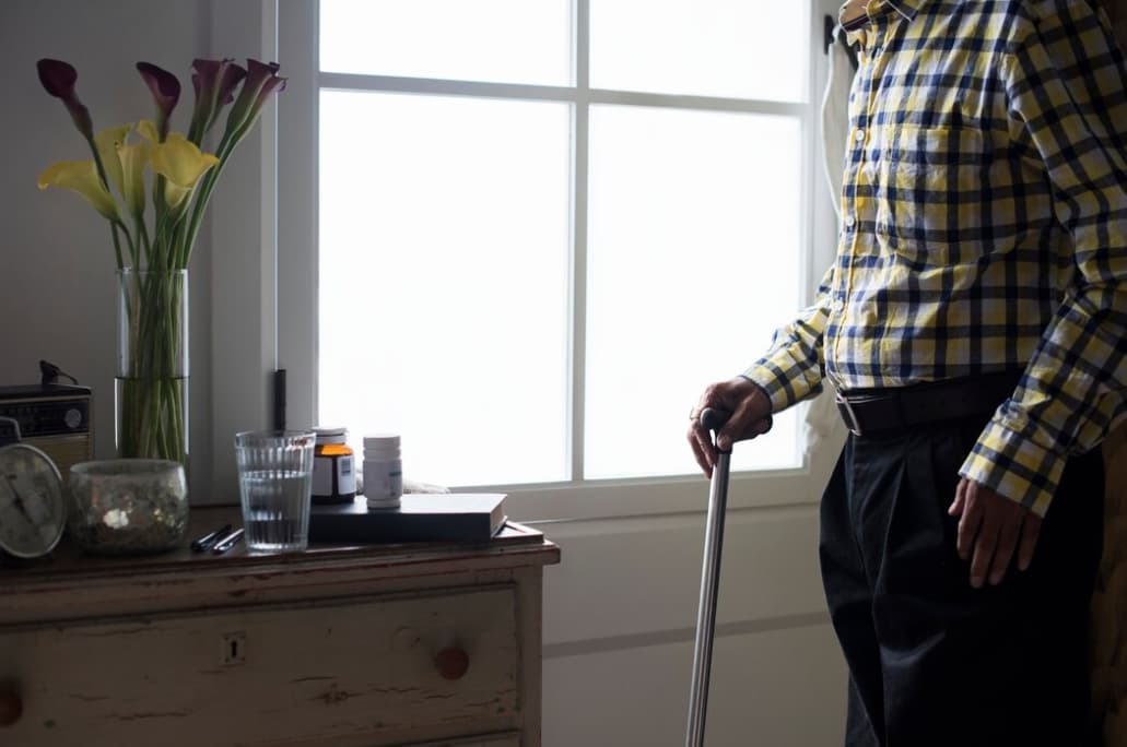A person standing with a cane beside a dresser with flowers and medication on top
